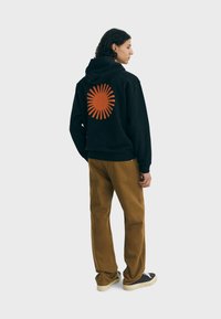 Felpa nera con un grande disegno di un sole arancione sulla schiena, abbinata a pantaloni marroni e sneakers nere, tessuto con texture, vestibilità comoda.