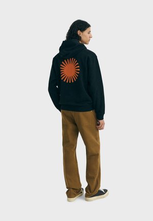 Sudadera negra con un gran diseño de sol naranja en la espalda, combinada con pantalones marrones y zapatillas negras, tejido texturizado, corte relajado.