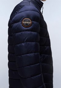 Tmavě modrá péřová bunda s vysokým límcem a obdélníkovou prošívanou strukturou. Má na rukávu kulatou nášivku značky.
