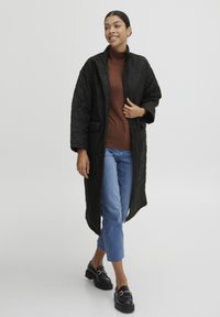 Abrigo largo acolchado negro con un alto cuello, con bolsillos laterales, llevado sobre un suéter de cuello alto marrón y combinado con jeans azules y mocasines negros.
