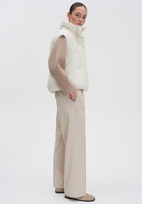 Puffige Creme-Weste mit hohem Kragen, kombiniert mit einem hellbraunen Langarmoberteil und beigefarbenen, weit geschnittenen Hosen, vor einem weißen Hintergrund.