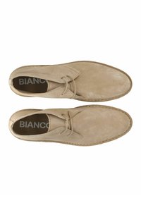 Zapatos de ante beige con punta redondeada, diseño de cordones y detalles de costura. El interior presenta la marca "BIANCO" sobre un fondo oscuro.
