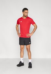 Punane lühikeste varrukatega spordisärk valge logoga, sobitatud mustade lühikeste pükste ja mustade tossudega. Lihtne disain, ilma mustrite või tekstuurideta.