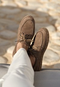 Brown suede shoes with laces, featuring an espadrille-style jute sole and metal eyelets, resting on a textured surface.
