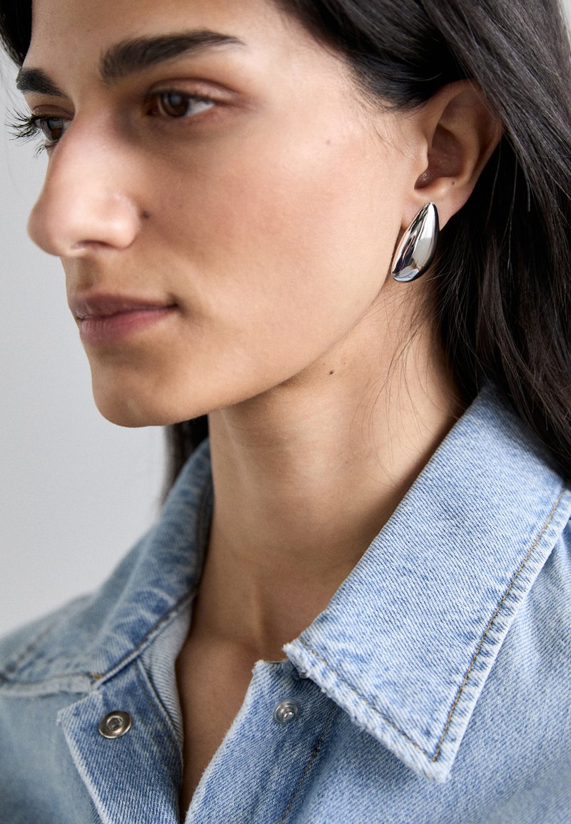 Srebrne uhane v obliki solzice so prikazane na modelovem ušesu, z poliranim zaključkom in gladkimi oblikami, nosi pa jih s svetlo modro jeans jakno.