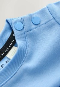 Világoskék pamut felső kerek nyakkal, két patenttal a vállnál, valamint márkanévvel ellátott címkével belül.