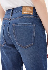 Närbild av en person som bär blå denimjeans med en läderetikett som står "ARMEDANGELS" ovanför bakfickan.