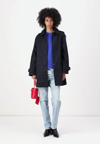Cappotto trench blu navy con chiusura a bottoni, caratterizzato da spalline sulle spalle e tasche laterali, abbinato a un top blu, jeans chiari e una borsa rossa.