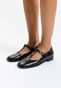 Mustat nahkaiset Mary Jane -kengät, joissa on T-nauha, neliskulmainen kärki, solkiyksityiskohta ja litteä korko. Silkkinen pinta, jossa on minimaalinen kiilto.