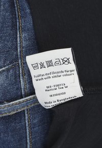 Hvidt plejeetiket på mørkt stof nær denim, med sorte vaske-symboler og tekst på flere sprog. Inkluderer mærke- og oprindelsesoplysninger.