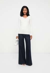 Maglietta bianca a maniche lunghe con scollatura a V e dettagli con logo, abbinata a jeans in denim scuro a gamba larga. Il modello tiene in mano una piccola borsa bianca.
