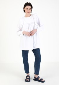 Weißes Langarmshirt mit Rüschendetails an den Schultern, kombiniert mit blauen Jeans und dunkelblauen Sandalen. Schlichte, lässige Silhouette.