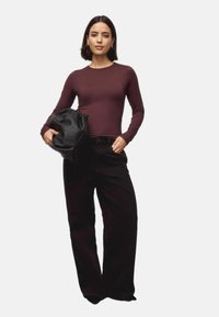 Frau in einem figurbetonten burgunderfarbenen Langarmoberteil und weit geschnittenen schwarzen Hosen, die eine große schwarze Leder-Clutch vor einem weißen Hintergrund hält.