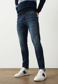Jeanși din denim albastru, cu o croială slim, ușor decolorati și cu un design cu cinci buzunare, asortați cu adidași albi cu dungi colorate.