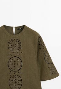 Olive green blouse with round neckline featuring intricate circular cut-out embroidery on front and sleeve, short wide sleeves, back button closure.