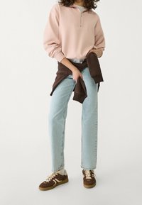 Blassrosa Zip-Up-Sweatshirt mit einer Vordertasche, kombiniert mit hellblauen geraden Jeans. Ein brauner Pullover um die Taille gebunden und braune Sneakers.