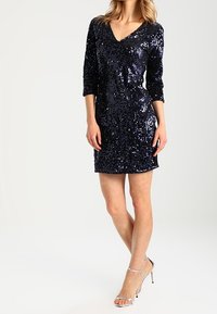 Abito blu navy con paillettes, scollo a V, maniche a tre quarti, design aderente, lunghezza corta, abbinato a tacchi argento con cinturini. Texture liscia.