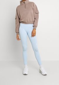 Brązowa bluza rozpinana na suwak z wysokim kołnierzem i bocznymi kieszeniami, w połączeniu z jasnoniebieskimi legginsami. Noszona z białymi sportowymi butami.