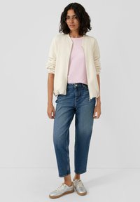 Veste bomber beige clair portée sur un t-shirt rose, associée à un jean bleu à jambes droites et des baskets argentées avec une semelle camel. Lunettes à monture transparente.