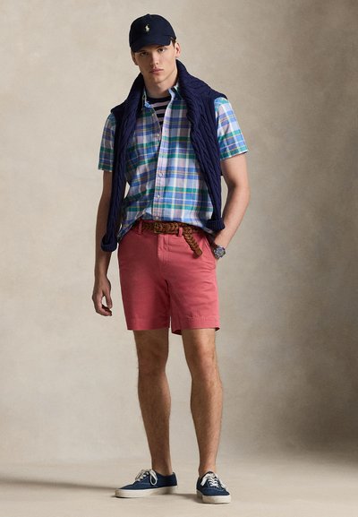 Πουκάμισο με κουμπιά και καρό σχέδιο σε μπλε, πράσινο και λευκό, σε συνδυασμό με κοραλί σορτς. Συνδυάζεται με μπλε πουλόβερ και καπέλο.