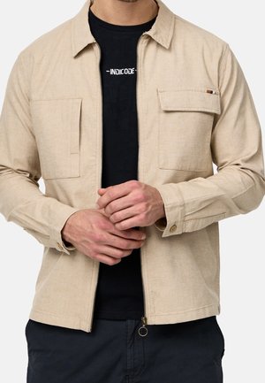 Homme portant une veste beige zippée sur un t-shirt noir avec un texte blanc, mains jointes devant, pantalon sombre visible à la taille.