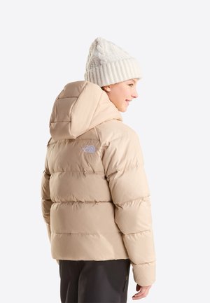 Personne portant une doudoune beige avec capuche et un bonnet blanc en tricot, vue de profil sur un fond clair uni.