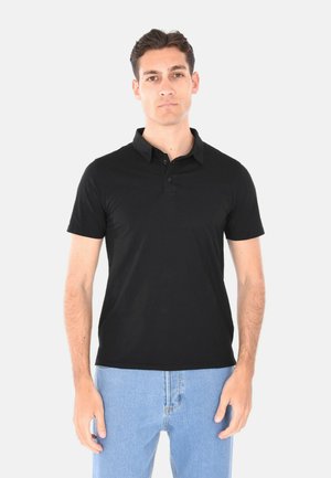 Uomo che indossa una polo a maniche corte nera e jeans azzurri chiari, in piedi davanti a uno sfondo grigio chiaro.