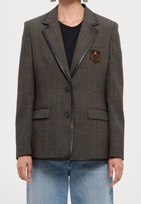 Šedý kostkovaný blazer s černým satinovým klopou, dvěma knoflíky, dvěma předními kapsami a zlatým emblémem na levé hrudi. Texturovaná látka.