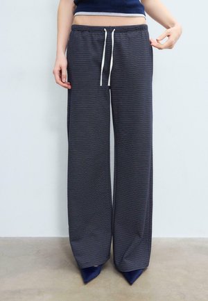 Osoba nosiaca voľné nohavice s námorníckymi modrými a bielymi pruhmi so štýlovými bielymi šnúrkami, v kombinácii so špicatými námorníckymi topánkami a krátkym námorníckym topom.