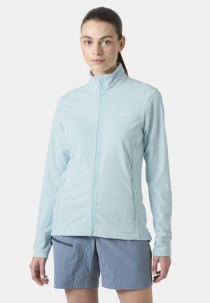 Mujer con cabello oscuro recogido en una coleta, vistiendo una chaqueta azul claro con cremallera y pantalones cortos grises, de pie frente a un fondo blanco liso.