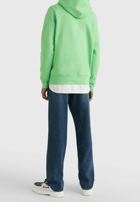 Felpa verde con cappuccio e polsini e vita a coste, indossata sopra una maglietta bianca. Combinata con pantaloni in denim blu e sneaker nere e bianche.