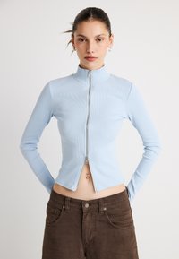 Γαλάζιο ριγωτό μπλουζάκι μακρυμάνικο με ψηλό γιακά και κεντρικό φερμουάρ· εφαρμοστή γραμμή και cropped μήκος. Καφέ παντελόνια φαίνονται από κάτω.