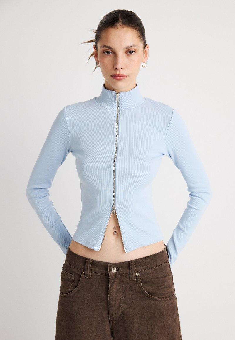 Γαλάζιο ριγωτό μπλουζάκι μακρυμάνικο με ψηλό γιακά και κεντρικό φερμουάρ· εφαρμοστή γραμμή και cropped μήκος. Καφέ παντελόνια φαίνονται από κάτω.