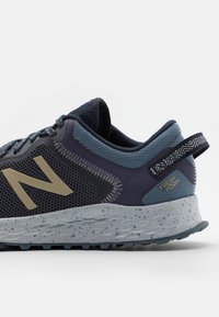 Zapatilla deportiva con parte superior de malla en azul oscuro y gris, con un gran logo beige, detalles texturizados y una suela gris moteada.
