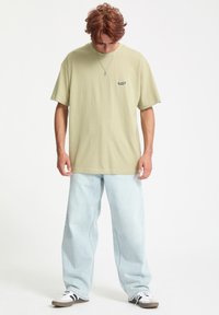 Oliwkowy bawełniany t-shirt z krótkim rękawem i małym czarnym logo, zestawiony z jasnoniebieskimi dżinsami o szerokich nogawkach oraz czarno-białymi trampkami.