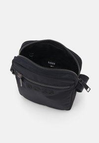 Borsa a tracolla nera in nylon con scomparto principale con zip, tasca frontale e logo impresso in modo sottile. Texture liscia con hardware resistente.