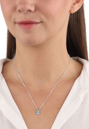 Collana con ciondolo in argento a forma di lucchetto, indossata su una camicia bianca, che mette in risalto il suo aspetto elegante e moderno.