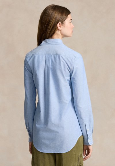 Naine näoga eemale, kandes hele sinist pika varrukaga särk nööbitud mansettidega ja oliivirohelisi pükse neutraalse tausta ees.