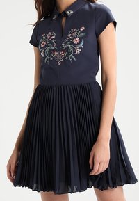 Marinblå klänning med krage, korta ärmar, blommönsterbroderi och plisserade kjoldelar. Nyckelhålöppning vid halsringningen.