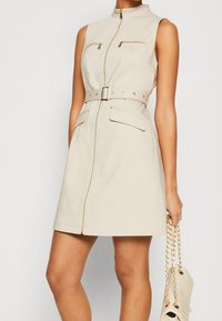Beige sleeveless dress with a high collar, front zipper, two chest pockets, belted waist, and a smooth texture. Carrying a cream handbag.