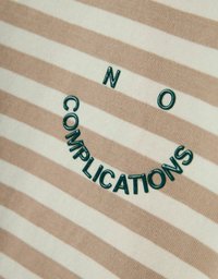 Tela a rayas en beige y crema; presenta texto bordado en verde formando una cara sonriente con la frase "SIN COMPLICACIONES."