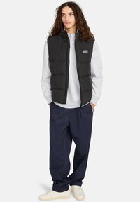 Schwarze gepolsterte Weste über einem hellgrauen Sweatshirt, kombiniert mit marineblauen Hosen und cremefarbenen Sneakern; Merkmale sind ein Reißverschluss und ein Logo-Detail.