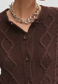 Cardigan en maille marron avec motifs en losanges texturés, doté d'une fermeture à boutons et de gros boutons marron. Porté avec un collier chaîne en argent.