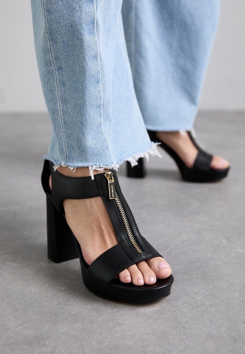 Pieds chaussés de sandales à talons noires avec une fermeture éclair dorée à l'avant, associées à un jean bleu clair à ourlets effilochés sur un sol gris.
