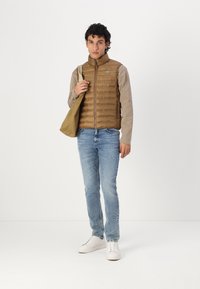 Vesta puf cu nuanță maronie, cusuturi orizontale, tricou cu mânecă lungă gri deschis, blugi albaștri deschiși și adidași albi. Ținând o geantă bej.