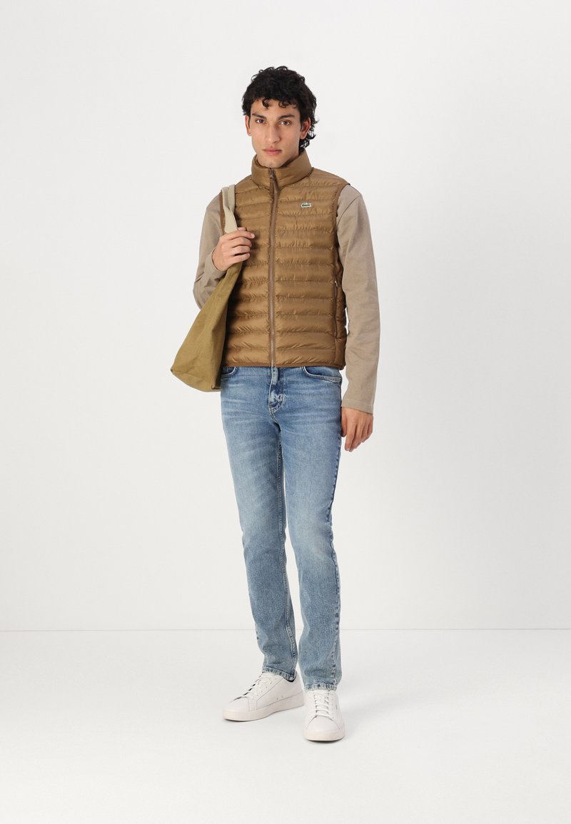 Braune Weste mit horizontalen Nähten, hellgraues Langarmshirt, hellblaue Jeans und weiße Sneakers. Hält eine sandfarbene Tasche.