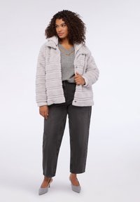 Sztuczna futrzana kurtka w jasnoszarym kolorze z prążkowaną teksturą, w zestawieniu z szarym swetrem i czarnymi spodniami z wysokim stanem. Buty to szare szpilki.