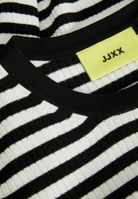 Tessuto a righe bianche e nere con una trama a coste testurizzata; presenta un colletto nero e un'etichetta gialla con la scritta "JJXX".