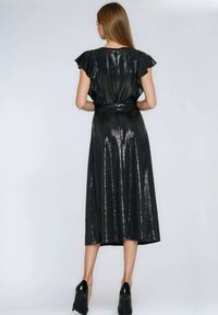 Kobieta ubrana w czarną, błyszczącą sukienkę z cekinów z krótkim rękawem i czarne szpilki, stoi tyłem do widza na jednolitym tle.