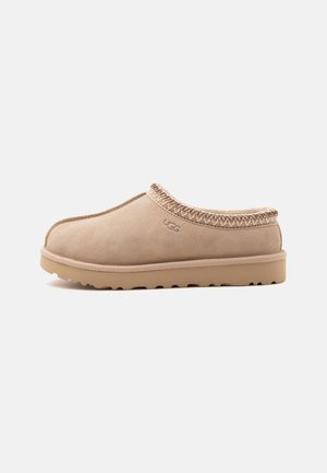 Chausson beige en daim à enfiler avec bordure cousue et semelle en caoutchouc, avec un petit logo UGG sur le côté.